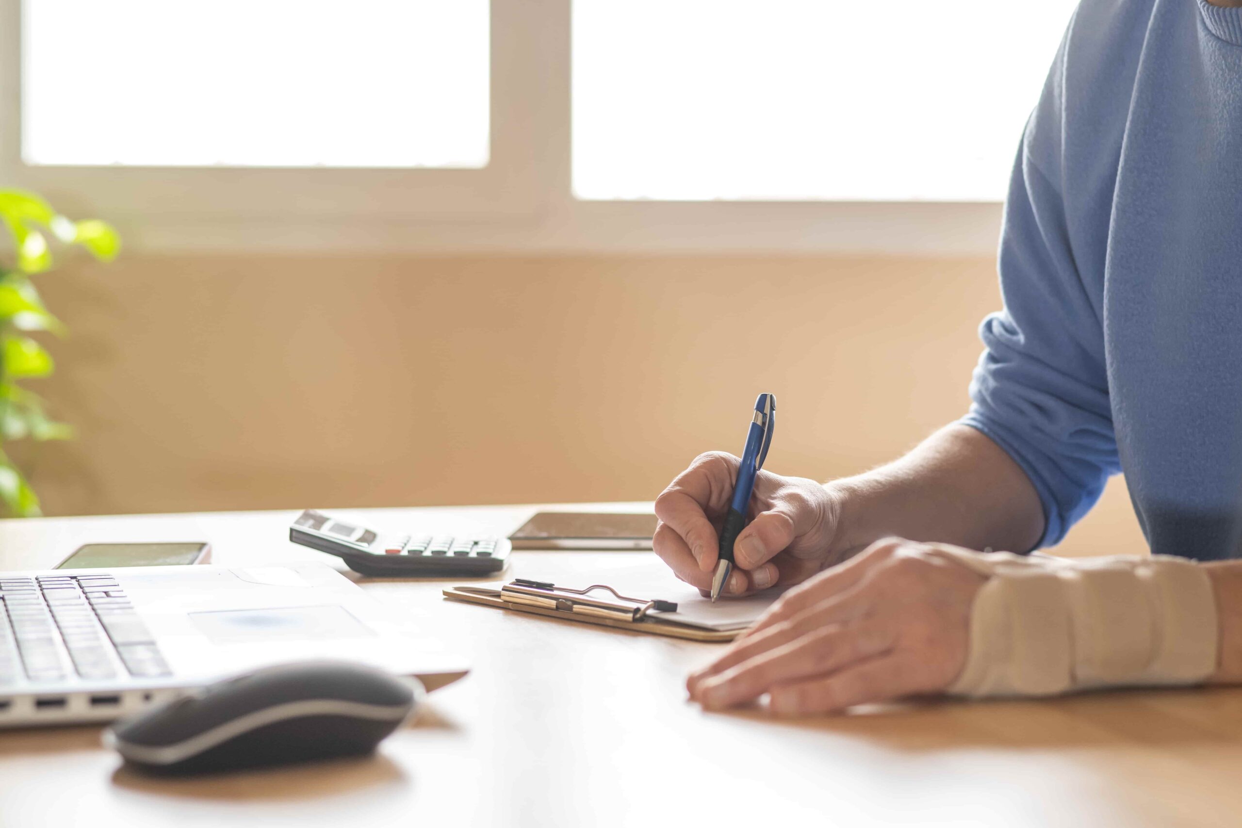 Injured person with a wrist brace filling out paperwork at a desk with a laptop, calculator, and documents, illustrating personal injury claim preparation.
