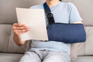 Injured man sitting on a couch reading a document with his arm in a sling, illustrating recovery after an accident.